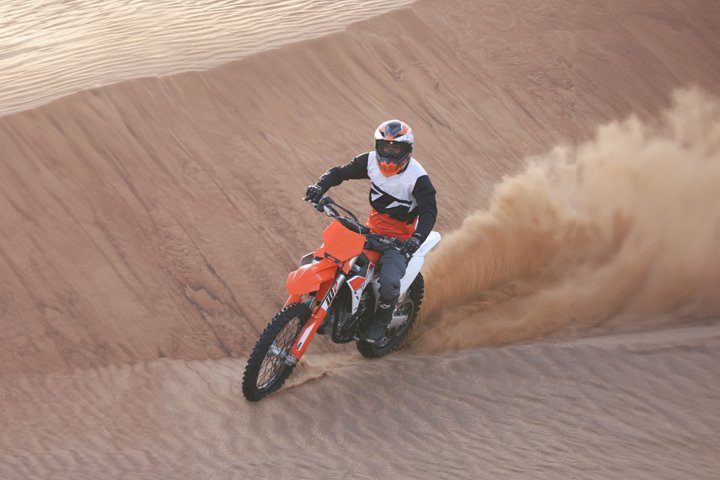 a guy riding dirt bike orange color ktm 450CC in the desert with sands swirling up from the back side